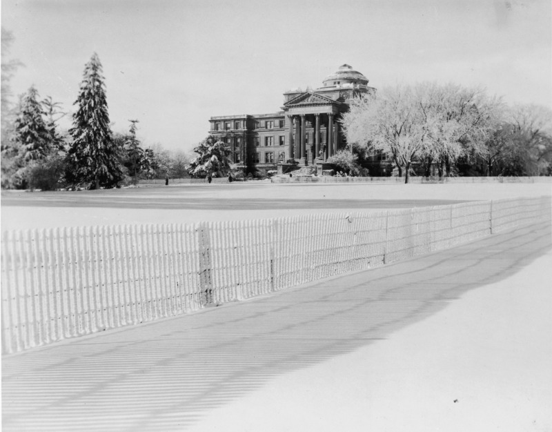 The front of Beardshear Hall from a snow covered central campus during winter. A snow fence is prominent. Four people are walking across central campus.