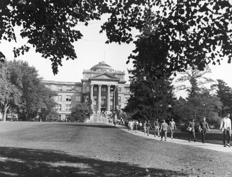 The front of Beardshear Hall from central campus. Students crowd the central sidewalk and front steps of Beardshear Hall. A utility truck is parked on the street. The image is reversed.