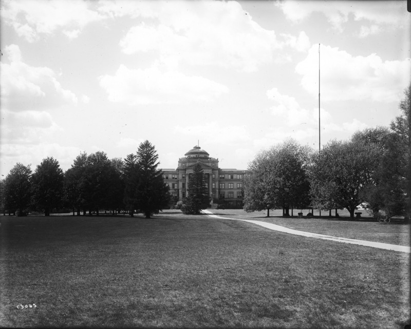 The front of Beardshear Hall from central campus. A central campus tree grove on the image right contains cannon and a flag pole.One vehicle is parked on the street in front of Beardshear Hall.