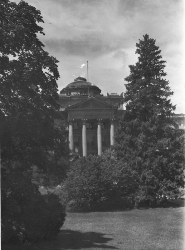 The portico and dome of Beardshear Hall as seen through shrubs and conifer trees from central campus.