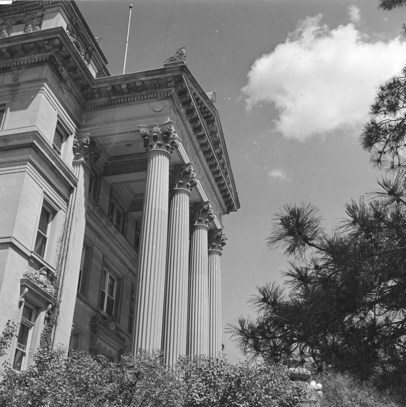 The portico of Beardshear Hall as seen from an extremely low angle. The image is framed by shrubbery and pine tree limbs.