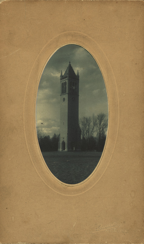 This closely cropped blue toned photograph depicts the Campanile in late fall or winter. The oval photograph is mounted on a formal presentation card embossed with "Lunde, Ames, Iowa.".