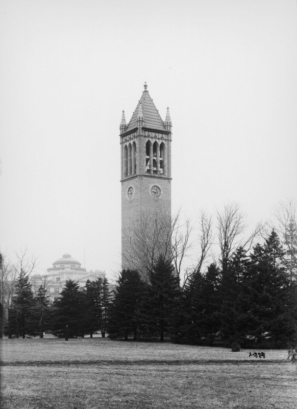 The Campanile in March 1906. Beardshear Hall is in the background on the image left behind a line of conifers. The same line of conifers obscures the view of the bottom of the Campanile. A path crosses the foreground from edge to edge of the image.