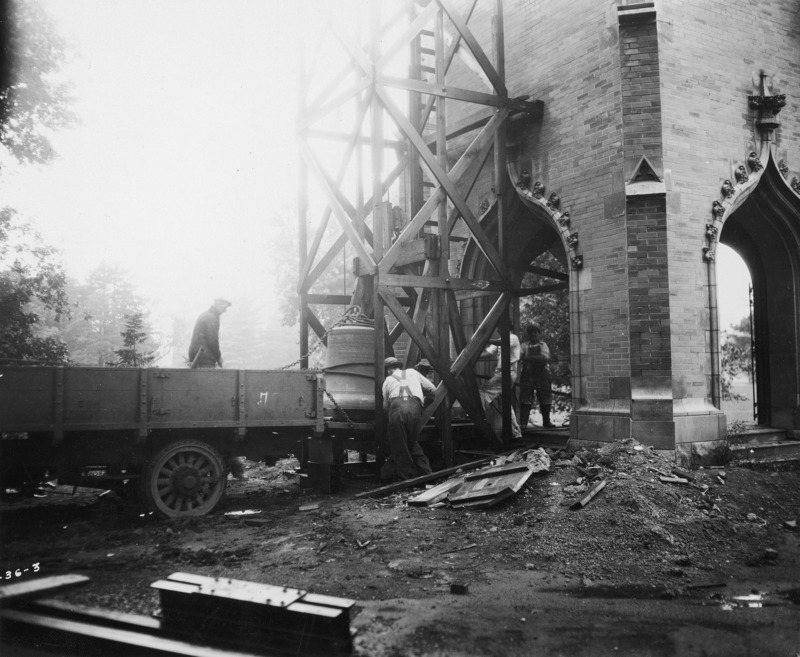 The base of the Campanile with a wooden tower built against one side. A delivery truck and some construction debris are at the bottom of the tower. The tower is for the purpose of installing a new bell in The Campanile. Four people are shown moving the bell from the delivery truck.