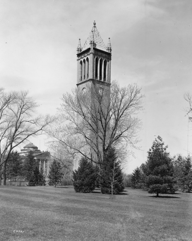 The east and south sides of the Campanile in early spring. The dome and portico of Beardshear Hall can be seen in the background through the trees. Foreground conifers obscure from view the base of the Campanile.