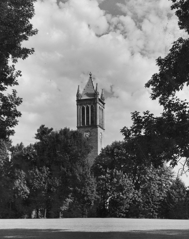 The Campanile in August 1945. The structure towers over a cluster of deciduous trees growing close to it. The clock shows 9:23. A long tree shadow covers the foreground grass.