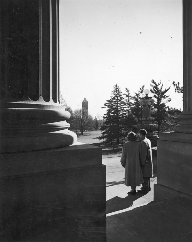 A couple on the steps of Beardshear Hall as seen from behind, framed by two giant columns. The Campanile is in the distance. The legend on the mount reads: "Students look from steps of Beardshear Hall toward Campanile." Each is wearing a coat.