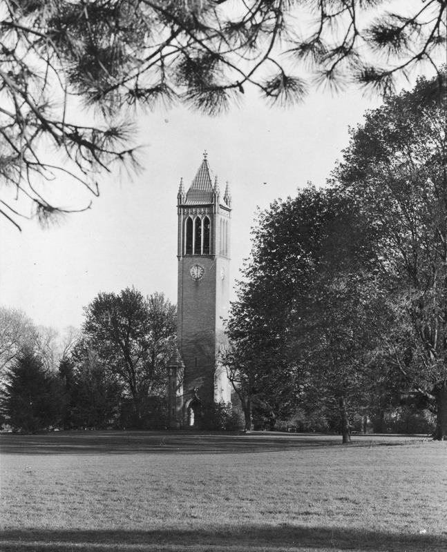 The Campanile as seen from the northwest. The structure sets in a landscape of shrubs and trees, with lawn opening up in the foreground. Overhanging bows of white pine frame the view. Clock shows 6:01.