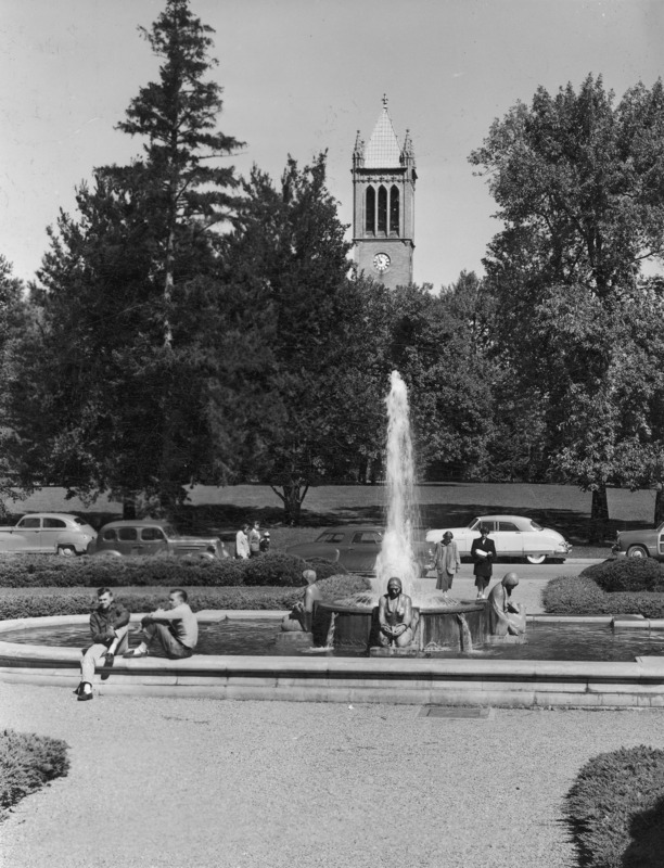 The Campanile rising above its surrounding trees, as seen from the north steps of the Memorial Union. Clock shows 10:47. In the middle distance is the Four Seasons fountain, which is running. Two male students are sitting on the near edge of the fountain. Behind the fountain people are walking on walkways. Automobiles are parked on the street.