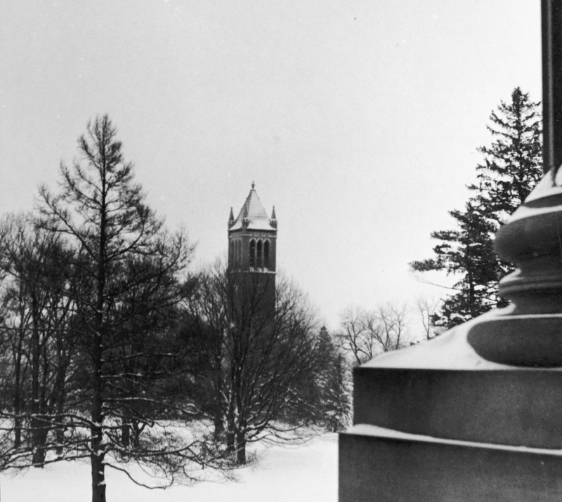 The Campanile in winter partially hidden among the trees on a snow covered central campus. The view is from the portico of Beardshear Hall, with the base of one of the large columns in the right of the image.