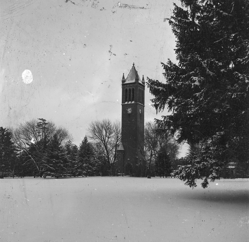 The Campanile on a snow covered central campus. Clock shows 3:11. A dark conifer on image right contrasts with the open expanse of central campus snow. There are surface stains on the photograph. One of a set of three photographs of the Campanile, each showing the same aspect of the tower but varying in location of the camera. The other photographs in the set are: 4-8-I.Campanile.230-7-4-1 and 4-8-I.Campanile.230-7-4-2.