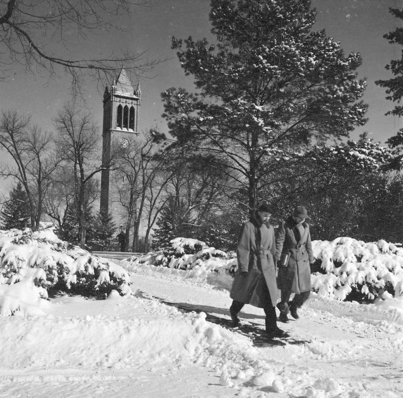 Central campus in winter. A snow covered sidewalk with two pedestrians is in the foreground with the Campanile in the distance. One of a set of five winter photographs, the others of which are: 4-8-I.Campanile.230-7-6-1; 4-8-I.Campanile.230-7-6-2 [not digitized]; 4-8-I.Campanile.230-7-6-3; and 4-8-I.Campanile.230-7-6-4.