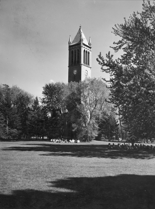 The Campanile rising out of the tree grove which surrounds the tower. Two classes are seated at two different sites on central campus, one directly in front of the Campanile and the other in the shade of a large tree on the left of the image. Clock shows 3:49.