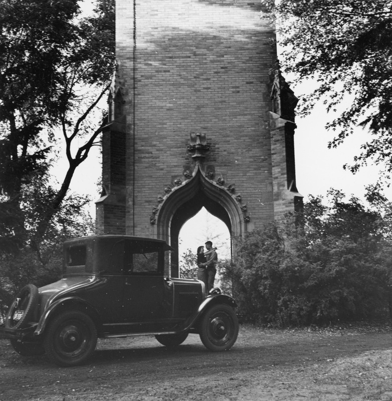 A couple, Wayne Brown (Veterinary Medicine degree, DVM1955) and Doris Blair Brown (Home Economics 1954), embracing under the Campanile. A car is parked in the foreground on a dirt road.