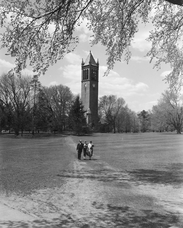 The Campanile in the distance with three people walking toward the camera in the foreground. Trees are just beginning to leaf out in this spring view. A well worn path runs from the camera to the Campanile.