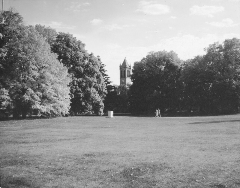 This view of the Campanile across a great open expanse of lawn shows full foliaged trees on either side of the view with two people in the middle distance walking across the lawn. An unidentified object (perhaps a weather data station) is in the middle distance.