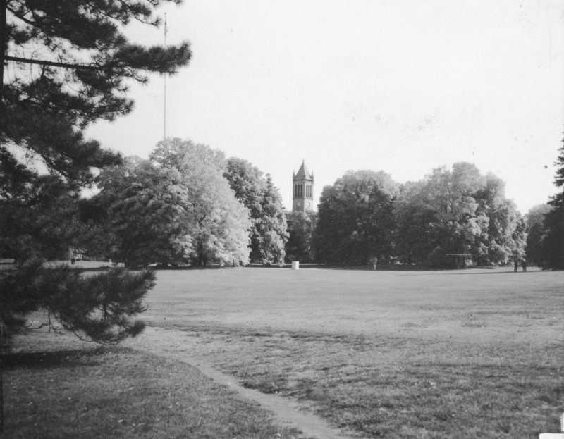 This view of the Campanile across a great open expanse of lawn shows full foliaged trees on either side of the view with one person walking along a path and two other groups to the left and right of the image. An unidentified object (perhaps a weather data station) is in the middle distance. A dirt path is in the immediate foreground.