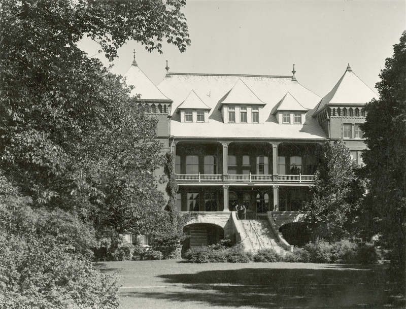 The front of Catt Hall with three men at the top of the stairs. The lawn and surrounding trees are in the foreground.