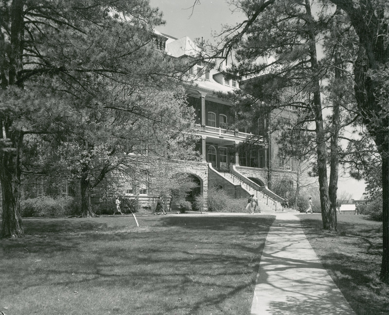 The front of Catt Hall with the lawn and sidewalk in the foreground. Several groups of individuals in warm weather clothing are walking on the sidewalk. The Greenhouses are barely visible in the background.