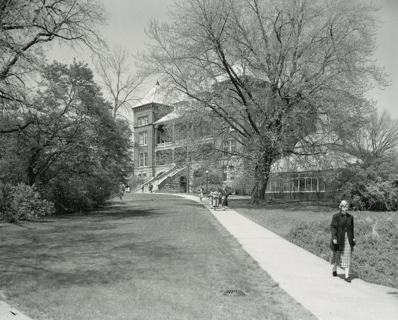 The front and side of Catt Hall. The greenhouse can also be seen through the trees. In the foreground, individuals can be seen walking along the sidewalk.