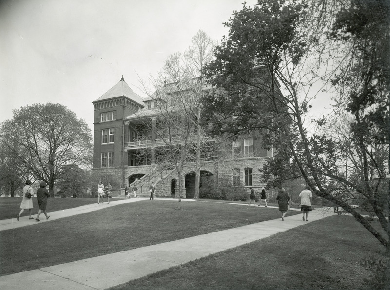 The front of Catt Hall. The lawn and surrounding foliage is featured in the foreground. Several individuals in period clothing are walking on the sidewalks in front of Catt Hall. This image is part of a set of images that includes: w 4-8-I.CattHall.234-4-3-2.