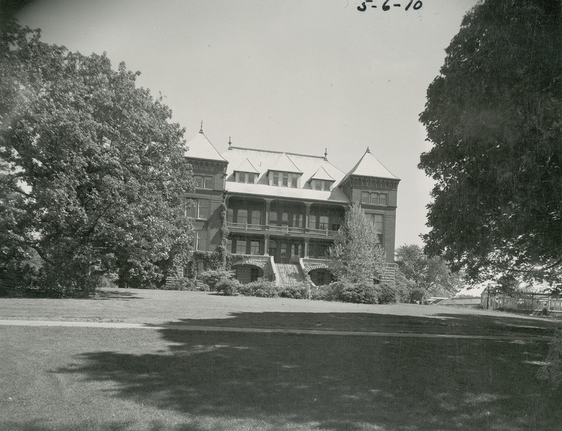 The front of Catt Hall. The building is partially obscured by the surrounding trees and bushes. The sidewalk and grass lawn are in the foreground of the image.