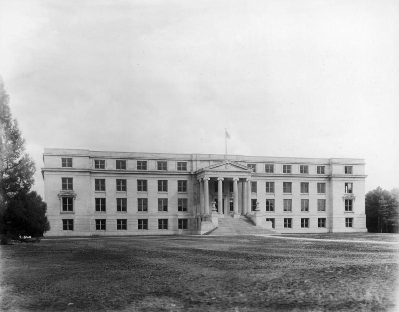 Curtiss Hall and the front entryway with its stairs, Ionic columns, and ornamental lamps. A flag is flying above the building. Trees are on either side of the building, and a treeless lawn stretches out in front of it.