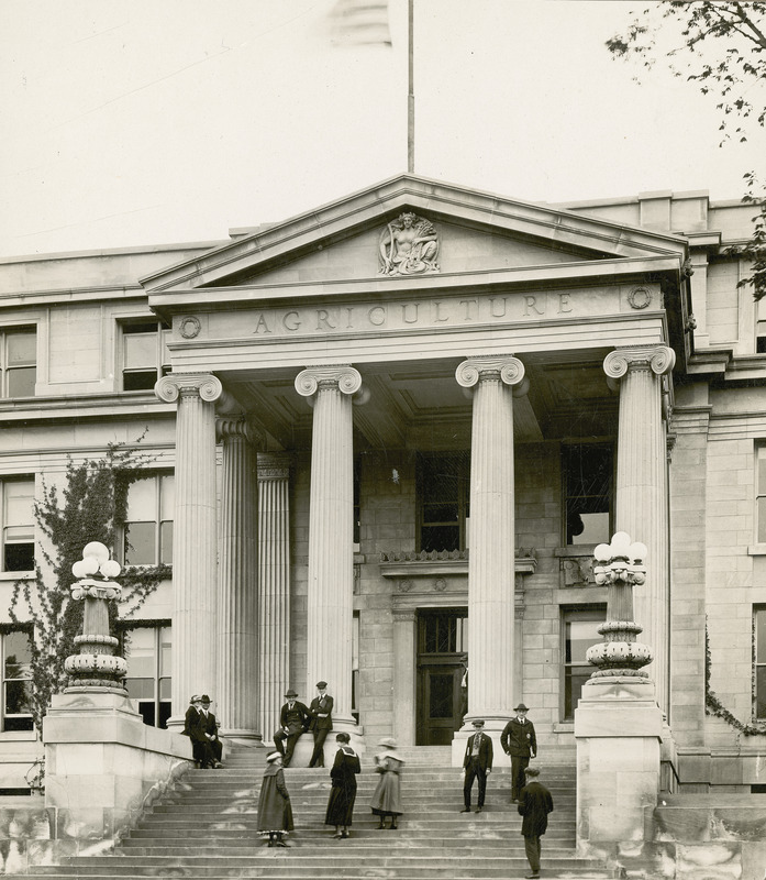 This photo shows the front entryway to Curtiss Hall with its Ionic columns, stairs, and ornamental lamps. Three men and three women are ascending or descending the stairs; four men and one woman are either sitting by or leaning up against the columns.