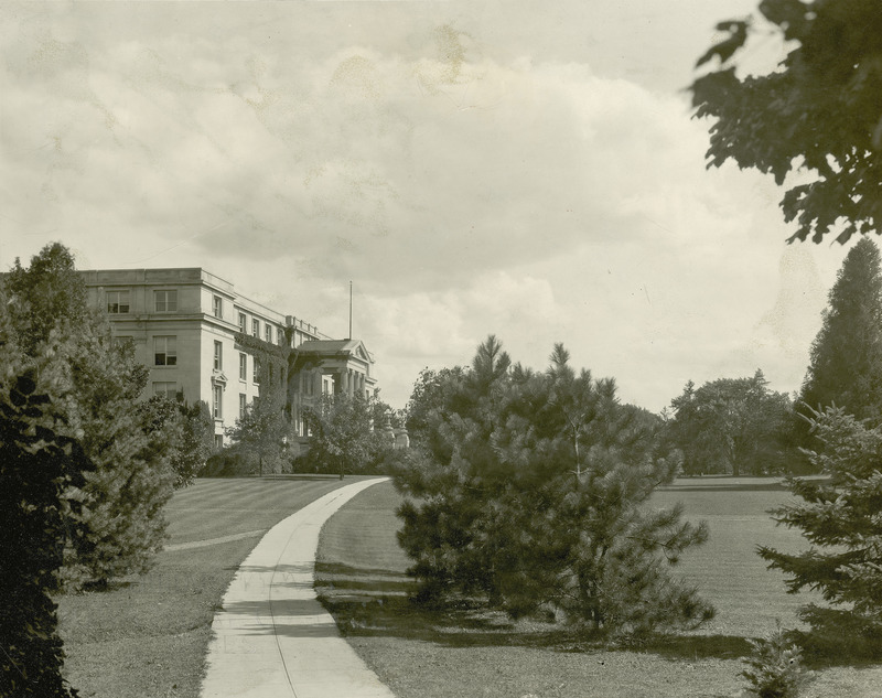 A curving sidewalk leads to Agriculture Hall (Curtiss Hall) from the north, showing young trees along the path and ivy beginning to grow on the building.