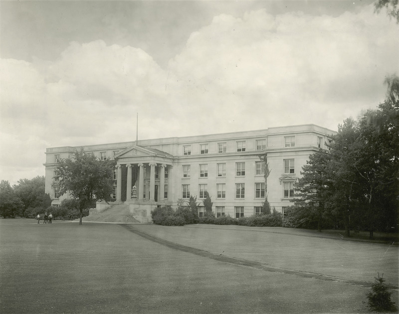 The front (west) facade of Agriculture Hall (Curtiss Hall) is viewed from the southwest, from a dirt path leading to the Campanile. Three students stand near the tree in front of the building.