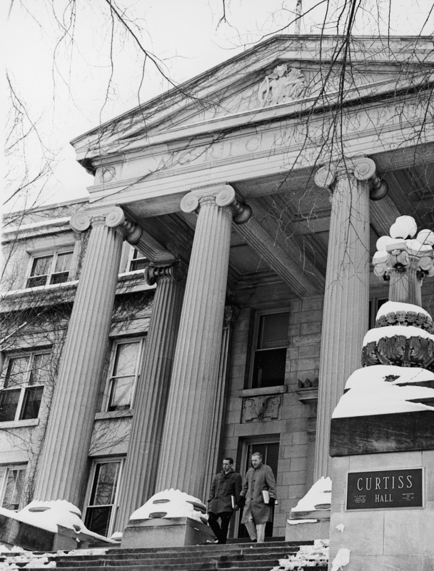 Two men in coats pass between the snow-covered bases of the columns as they leave Curtiss Hall. The name plaque on the building indicates it was presented by the class of 1957.