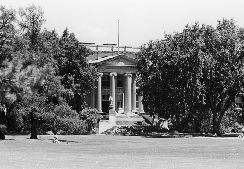 Curtiss Hall is viewed from central campus with bicycles and a fence in front and students sitting under trees on either side.