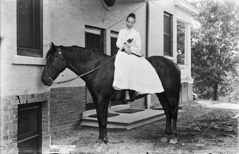 An unidentified woman wearing a white dress and holding a cat, sits sidesaddle on a saddle horse near one of the entrances to the Knoll. A good view of the building's brick foundation appears.