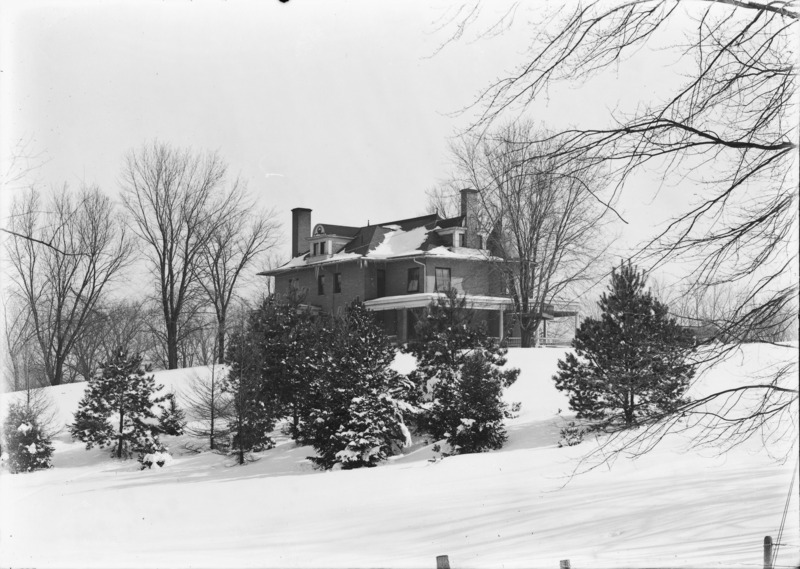 This winter view of the Knoll focuses on the southwest corner. Trees and bushes are in the foreground. There is snow on the roof and the surrounding lawn.