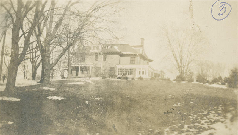 This westerly view of the Knoll features the large glassed-in porch on the right and the porte cochere on the right. There are trees and grass in the foreground.