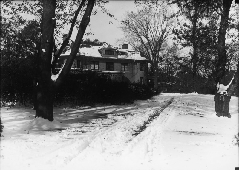 The eastern side of the Knoll is partially screened from view by the trees and shrubbery. The porte cochere and a garage or storage building can be seen in the distance. The roof, trees and lawn are covered with snow.