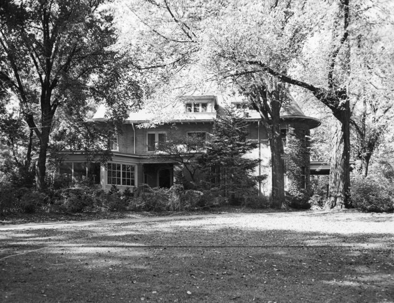 The east side of the Knoll is almost hidden from view by the trees and other shrubbery. The large wrap-around porch on the left is either glassed or screened in. The porte cochere is partially visible through the trees on the right.