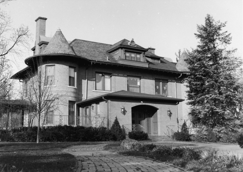 This view of the eastern side of the Knoll shows the front entryway and open air porch. A bricked sidewalk runs in front of the house. The right tower is hidden by a large tree.