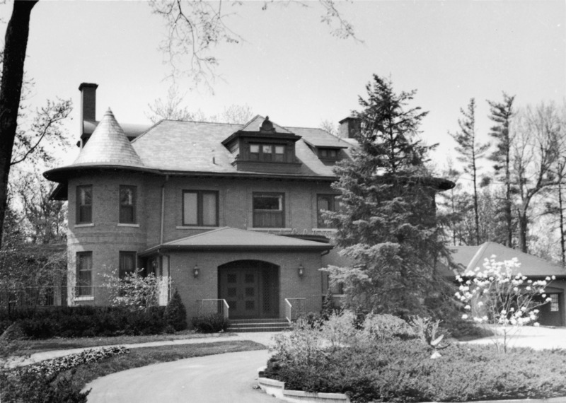 The front of the Knoll. The home's dormers, towers, chimneys, entryway, driveway, and garage are visible. An evergreen partially blocks the view of one tower; and a small, blossoming tree partially blocks that of the garage.