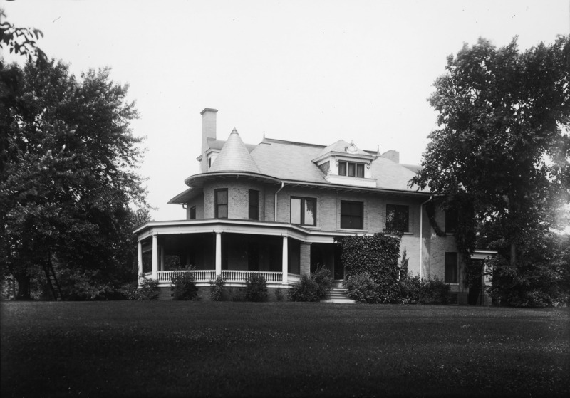 The eastern side of the Knoll is surrounded by trees in this ground level view. The large wrap around porch is visible on the left and the entryway is covered with ivy. The porte cochere is nearly hidden by trees.