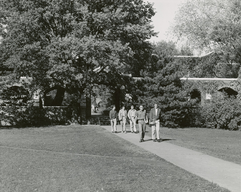 Five men are walking down the sidewalk in front of the south entrance to the Quadrangle (later known as Lagomarcino Hall).