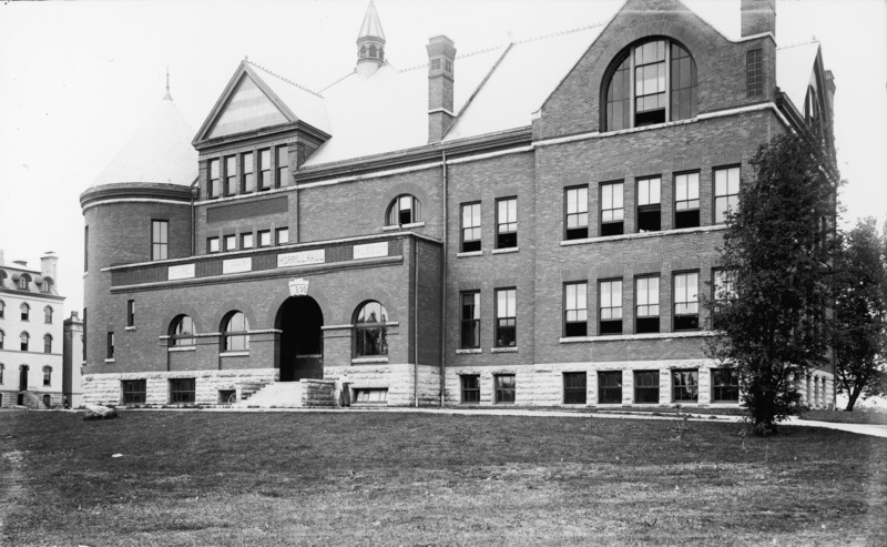 View of the front (east) side of Morrill Hall. A woman walking northwards is beside the main entryway to the building. Old Main is on the left side of the photo.