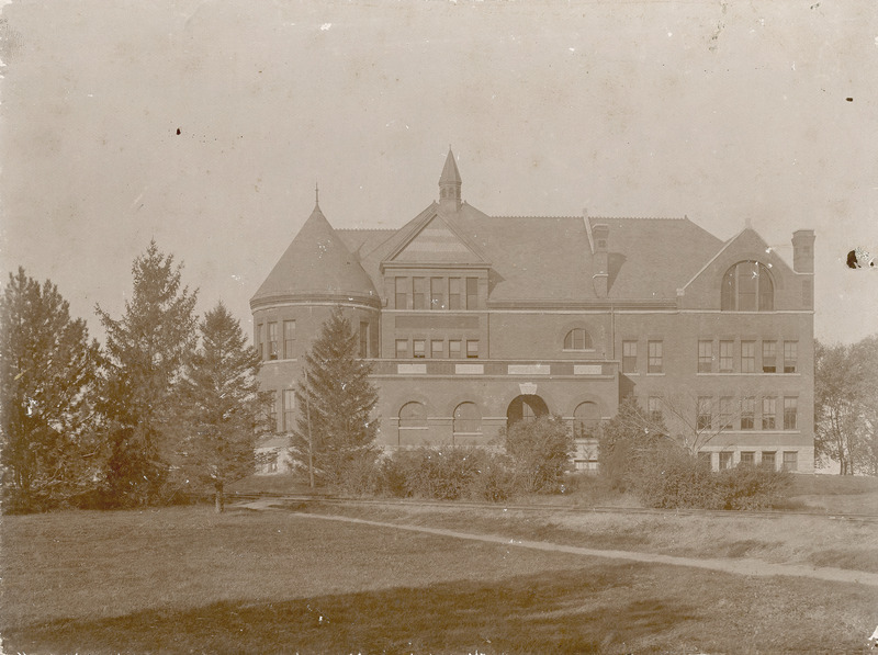 The front (east) side of Morrill Hall is partially obscured by trees and scrubs in this view. The tracks of the Ames and College Railway (the Dinkey) are seen in front of the foliage.