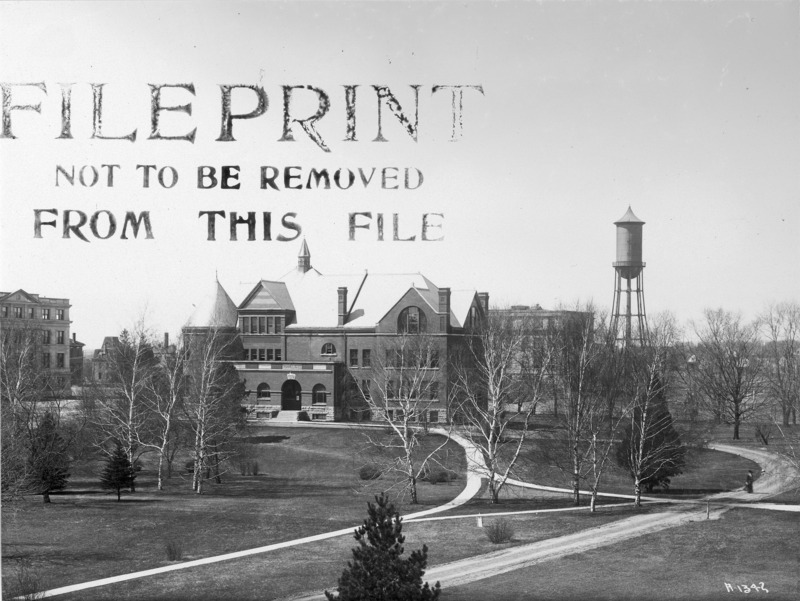 Slightly elevated view of the front (east) and north sides of Morrill Hall with a long sidewalk leading up to the building. A woman is on a dirt road to the north of the building. Marston Hall and water tower are in the back right of the photo. Part of Beardshear Hall is on the left of the photo.