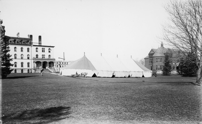 A large tent is pictured in the center of this photograph. Old Main is to the left of the tent and Morrill Hall is on the right. Marston Hall (under construction) can be seen in the distance. A man in a horse-drawn wagon is in front of the tent.