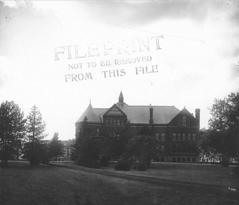 The front (east) of Morrill Hall is partially obscured from view by trees. Marston Hall is visible in the background and the tracks for the Ames and College Railway (the Dinkey) are visible in the foreground.