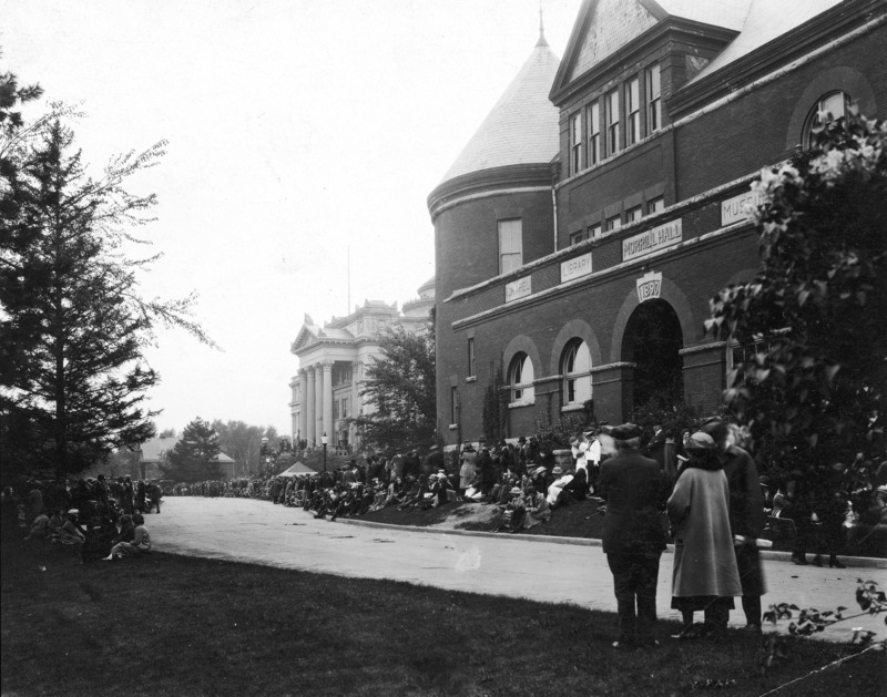 A crowd is gathered along Morrill Road in front of Morrill Hall and Central Building (Beardshear Hall). A small tent appears between the two buildings. A period automobile is visible near the English Office Building in the background.