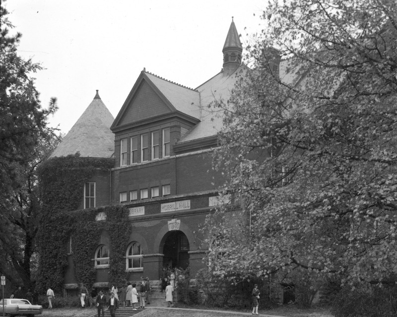 Students are gathered around the front entrance to Morrill Hall. Ivy is growing on the outside of the building. A period automobile is parked in front of the building.