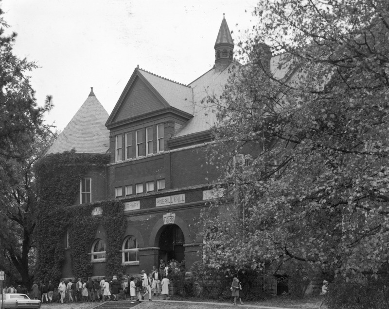 Students are gathered around the front entrance to Morrill Hall. Ivy is growing on the outside of the building. A period automobile is parked in front of the building.