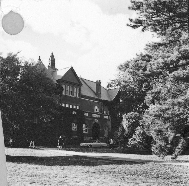 View of the front of Morrill Hall, partially obscured by trees. Several students are walking in front of the building. A car is parked in front of the building.